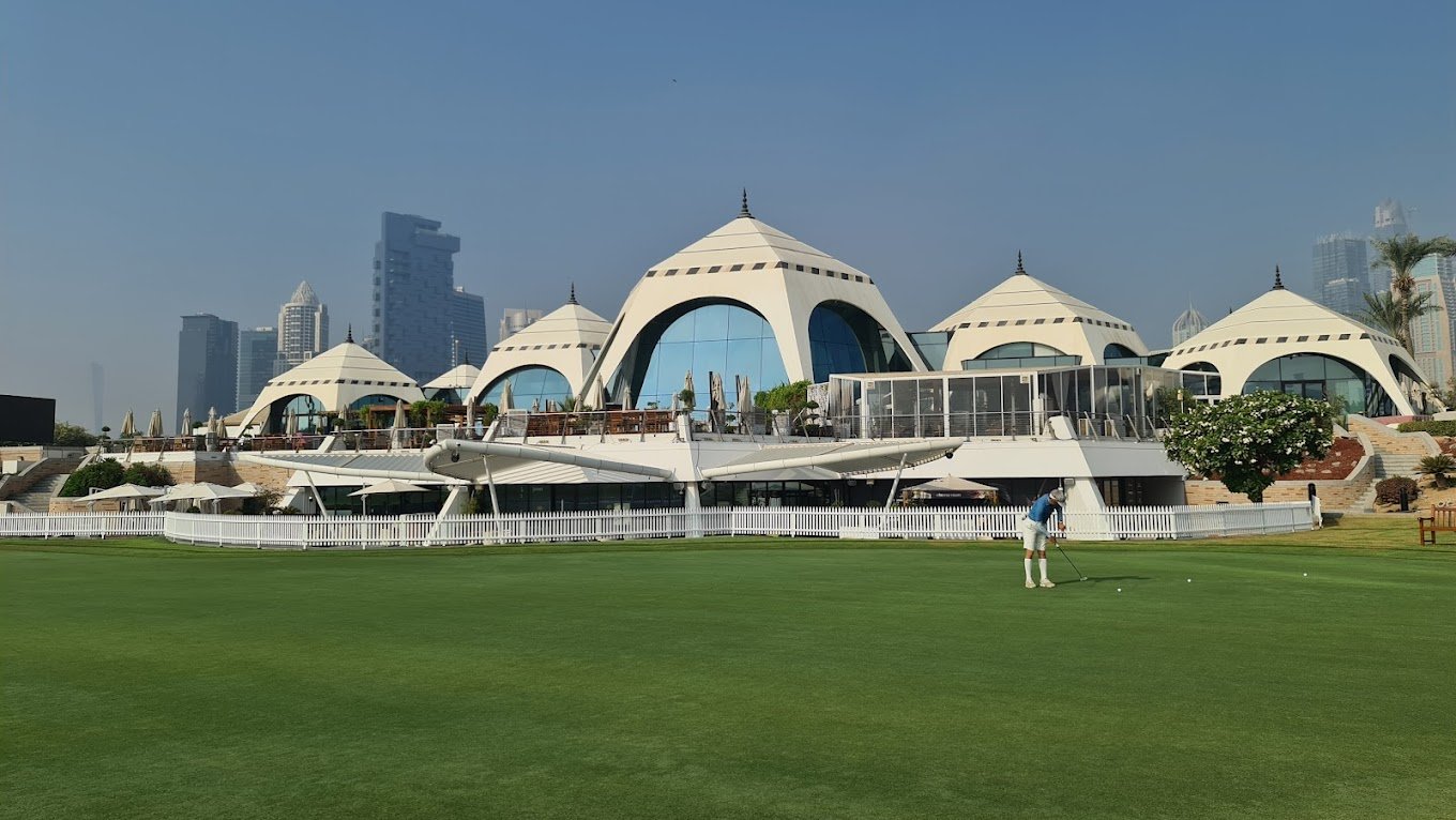 Scenic view of Emirates Golf Club featuring a lush green golf course with palm trees, a modern clubhouse, and the Dubai skyline in the background under a clear blue sky