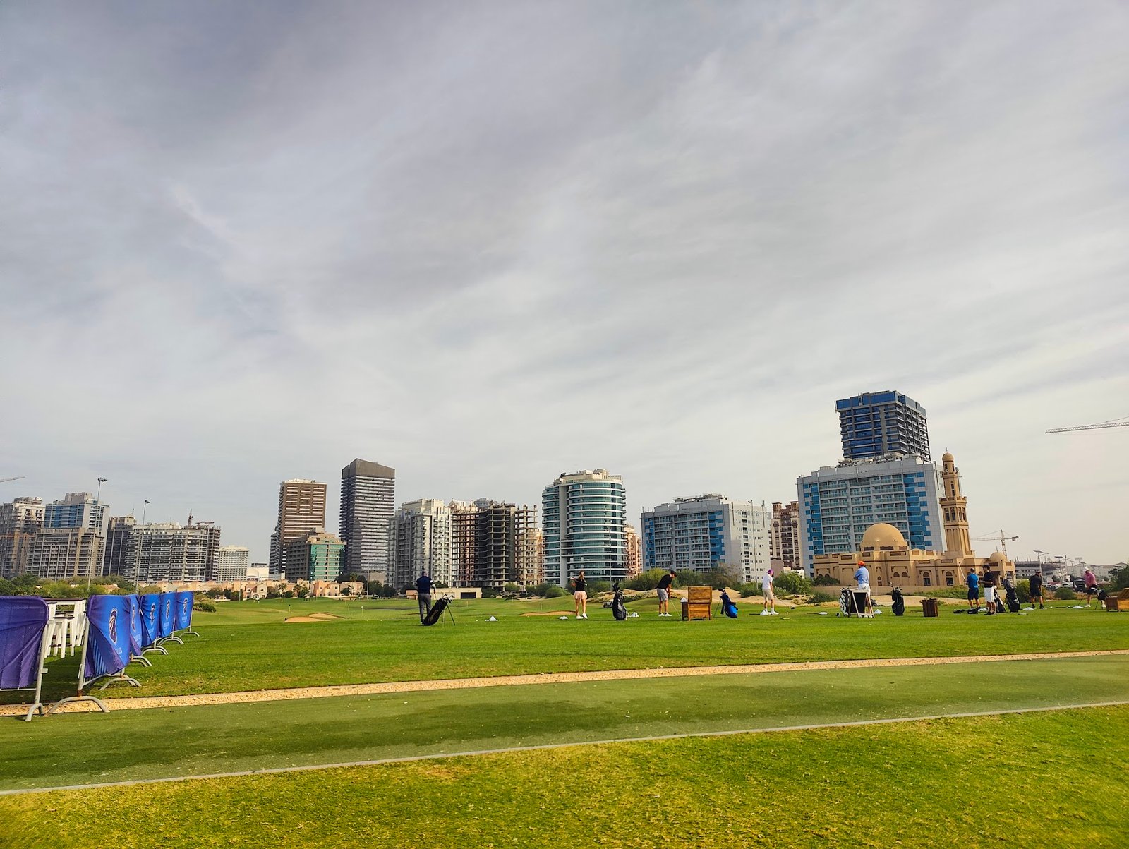 Wide view of The Els Club golf course with rolling green fairways, sand bunkers, and a modern clubhouse in the distance under a clear blue sky.