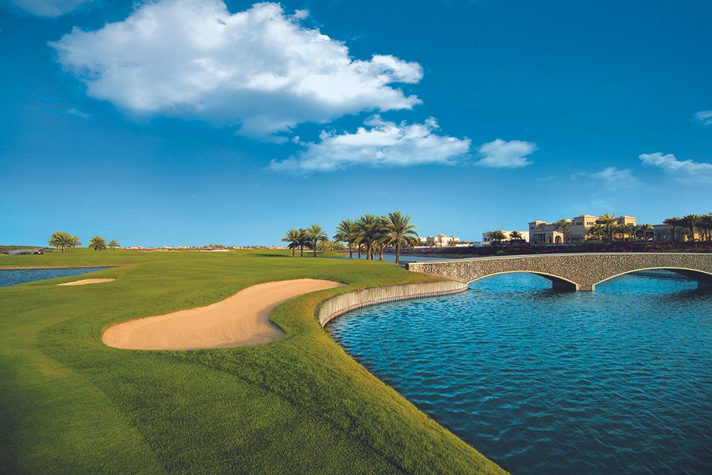 Desert-style Arabian Ranches Golf Club featuring sandy landscapes, green fairways, and a traditional-style clubhouse under a clear blue sky.